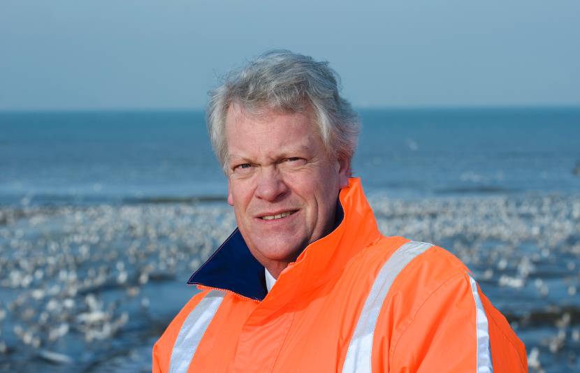 Portret deltacommissaris in oranje windjack met zee op achtergrond groot